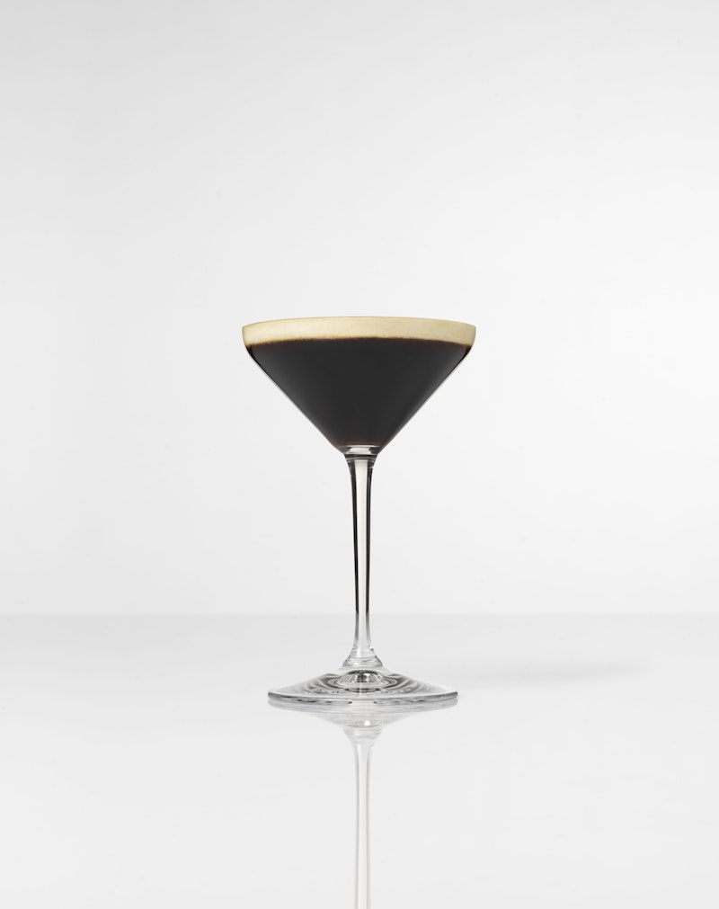 Cocktail glass filled with Espresso Martini, with white foam on top, on a white background.